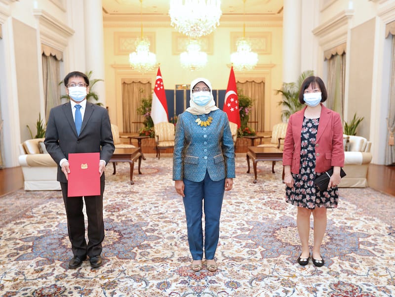 Three people wearing masks stand in a room with Singapore flags.